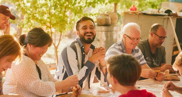 Guests laugh together during an outdoor communal meal under leafy trees.