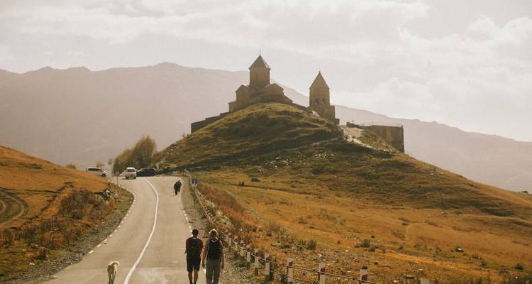 Hikers walk a country road toward a hilltop church set against hazy mountains.