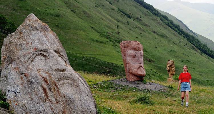 Traveller walks past large carved stone faces on a green mountainside meadow.