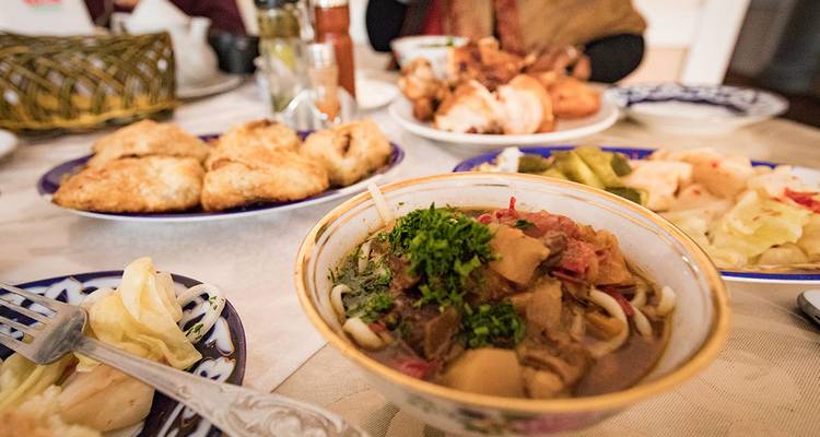 Plateau de repas ouzbek présentant une soupe copieuse, des pâtisseries et des légumes marinés sur des plats décorés.