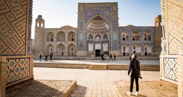 Un voyageur admire la façade en mosaïque complexe d'une grande madrasa par une belle matinée ensoleillée, encadrée par des portails en arche.