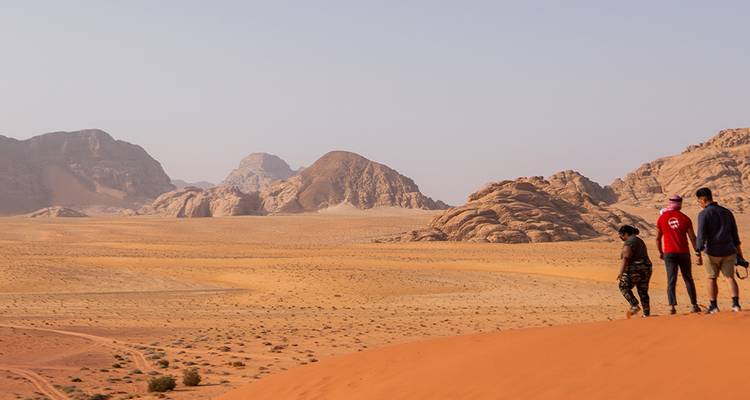 Three hikers walk along an orange sand dune with rugged Wadi Rum mountains in the distance.