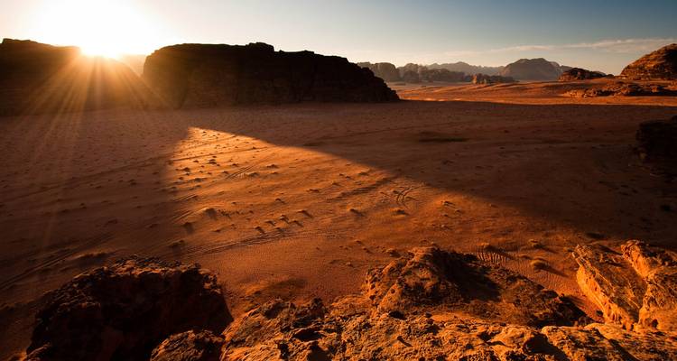 Golden sun rays cast long shadows across the dramatic sandstone landscape of Wadi Rum at sunrise.