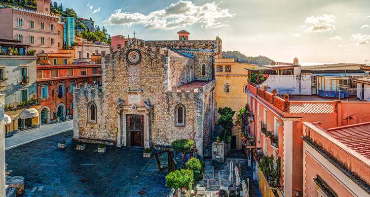 Plaza histórica llena de color en Taormina con una iglesia de piedra y vistas al mar bajo la luz del atardecer.