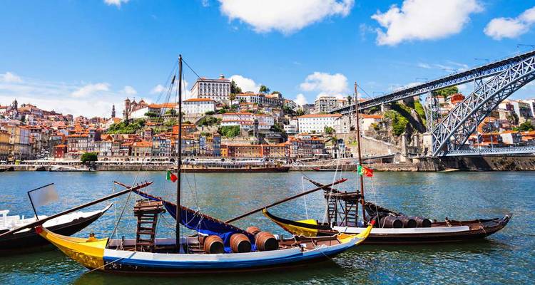 Traditional rabelo boats loaded with wine barrels moored on the Douro River with Porto’s colourful hillside skyline and iron bridge behind.