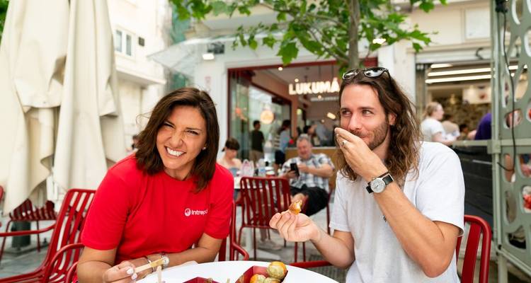 Two travellers laugh while sampling Greek pastries at an outdoor café in Athens.