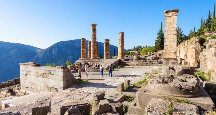 Visitors explore the ruined Temple of Apollo at Delphi set against forested mountains and blue sky.