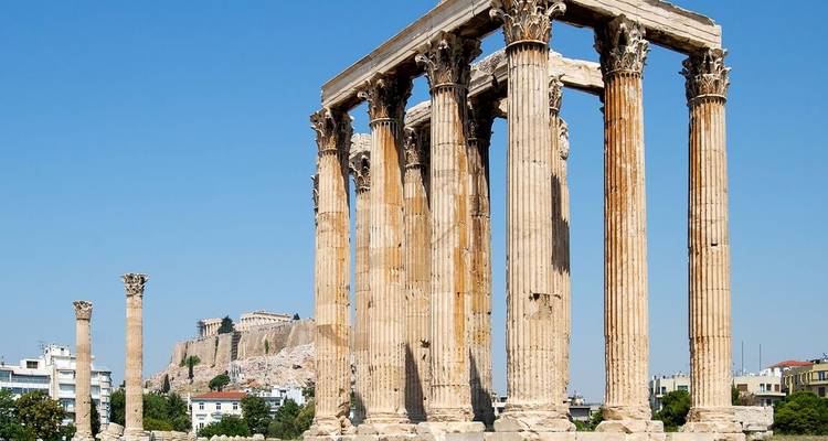 The towering Corinthian columns of the Temple of Olympian Zeus with the Acropolis rising in the background under a bright blue sky.
