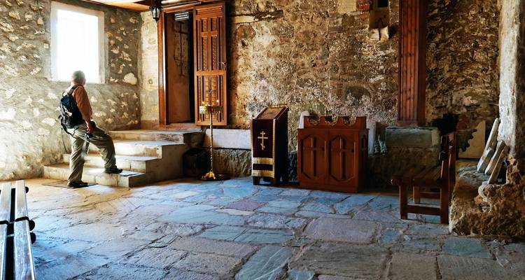 A lone visitor with a backpack pauses inside a dim, stone-walled chapel illuminated by morning light from a small window.
