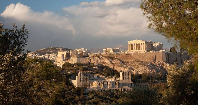 A golden-hour panorama of the Acropolis hill framed by pine trees, the Parthenon glowing above Athens.