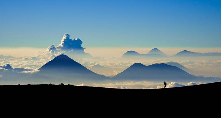 Silhouetted trekker against dramatic cloud layers and distant volcanic peaks at sunrise.