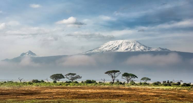 Snow-capped Mount Kilimanjaro rising above acacia-dotted plains under a partly cloudy sky.
