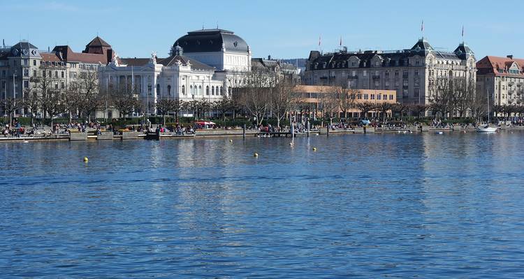 Frente urbano junto al agua con teatros históricos y hoteles reflejándose en un lago tranquilo.