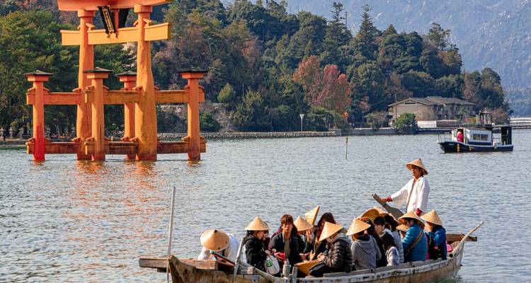 Un tradicional barco de madera con turistas pasa por el icónico torii flotante en una bahía tranquila.