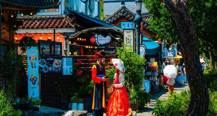 Pareja en hanbok tradicional pasea por una colorida calle de pueblo hanok decorada con linternas y tiendas.