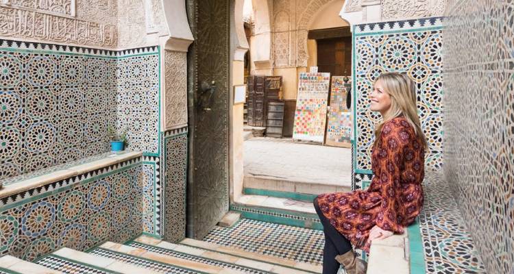 A female traveller sits on mosaic-tiled steps inside an ornate medersa adorned with intricate stucco and zellige.