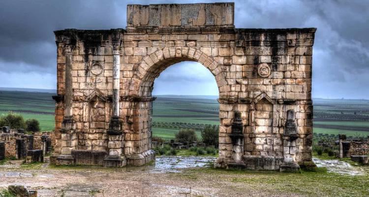 The triumphal arch of Volubilis stands weather-worn beneath brooding grey skies and lush farmland.