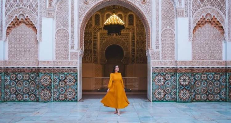 A woman in a flowing yellow dress strolls through a grand Moroccan courtyard adorned with elaborate arches and mosaic tiles.