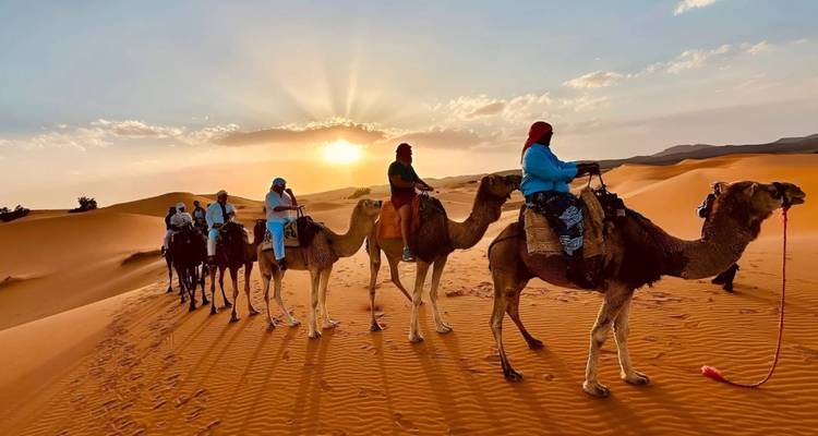 At sunset, travellers ride camels across glowing orange dunes as the sun dips behind the horizon.