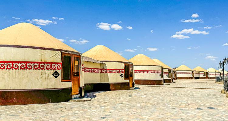 A neat row of cream-colored yurts with red patterns sits on a wide stone courtyard under a blue sky.