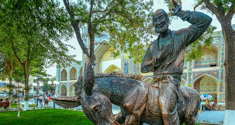 A whimsical bronze statue of a man riding a donkey stands beneath leafy trees near an ornate mosque.
