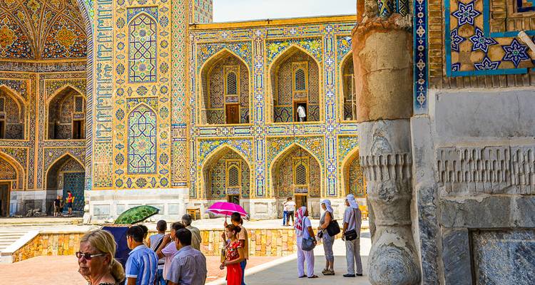 Tourists with colorful umbrellas explore the intricately tiled facades of Registan Square.