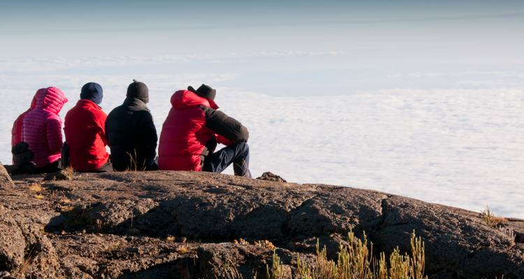Quatre trekkeurs vêtus de vestes rouges sont assis sur une saillie rocheuse et contemplent une mer de nuages.