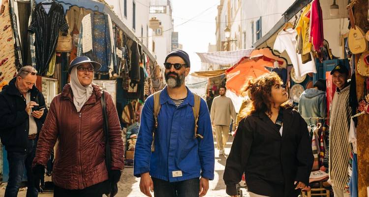 Les voyageurs se promènent dans une rue de marché ensoleillée bordée d'étals et de textiles.