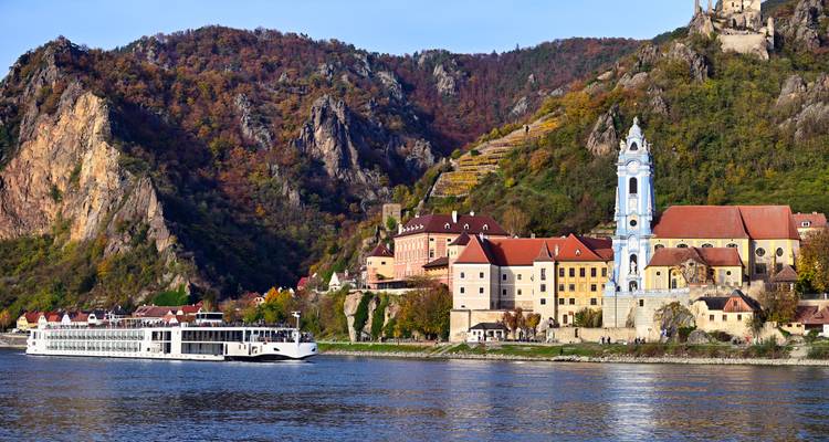 Elegant white river cruise ship glides along the Danube past the blue-towered abbey and steep vineyard-covered cliffs of a colorful medieval village.