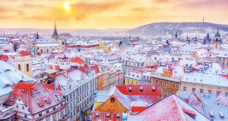 Snow-covered rooftops of Prague’s Old Town glow pink under a winter sunrise, with church spires piercing a pastel sky.