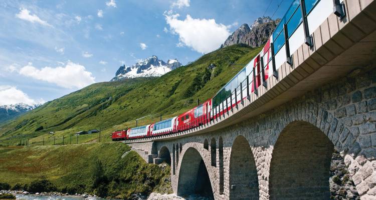 Red panoramic train crosses a curved stone viaduct in the lush Swiss Alps with a snow-capped peak and blue sky behind.