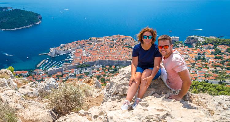 Ein Paar posiert auf dem felsigen Berg Srđ mit Panoramablick auf Dubrovniks ummauerte Stadt und das tiefblaue Meer darunter.
