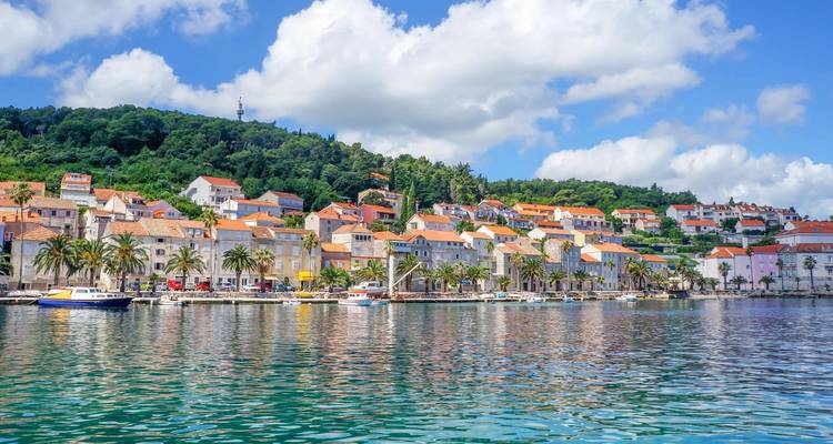 Colorful seaside village with terracotta rooftops and palm-lined waterfront reflected in clear Adriatic water.