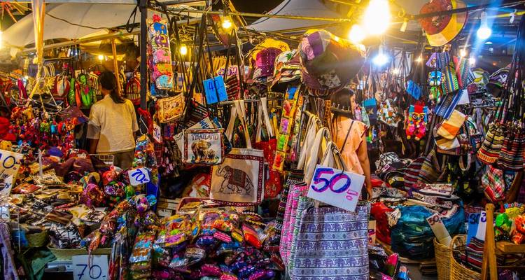 Bustling Thai night market stall overflowing with colorful textiles, bags and trinkets lit by warm hanging bulbs.