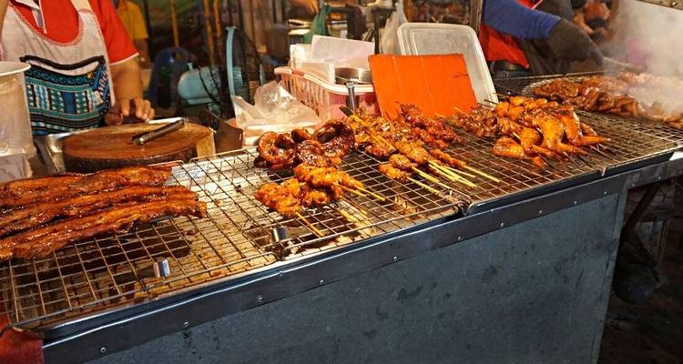 Street food vendors grill skewers of glazed meat over hot coals at a lively night market food stall.