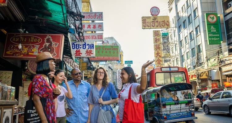 Small tour group chats on a busy Bangkok street lined with colourful shop signs, tuk-tuk and overhead cables.