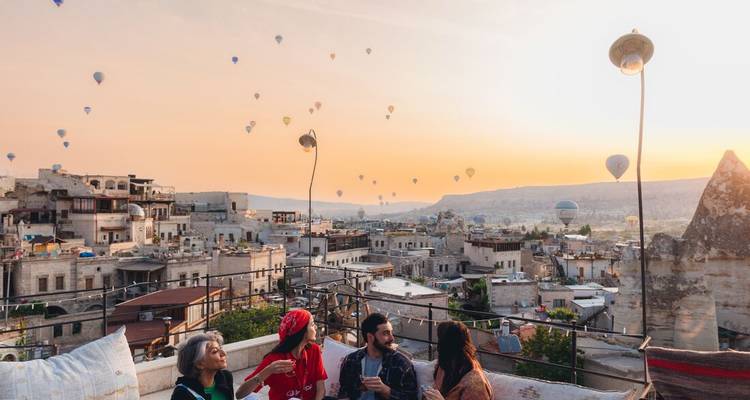 Amigos relajándose en una terraza en la azotea al atardecer, con vista a las casas de piedra de Capadocia mientras globos aerostáticos flotan en el cielo.