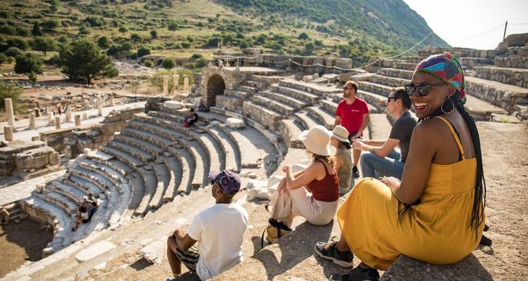 Los viajeros se sientan en las antiguas gradas de piedra de un anfiteatro grecorromano rodeado de colinas verdes.