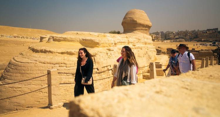 Un pequeño grupo de turistas camina pasando la Gran Esfinge en un día soleado del desierto con el horizonte de la ciudad débilmente visible más allá.