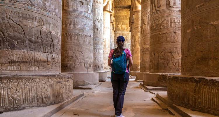 Un viajero con mochila camina entre enormes columnas de piedra tallada cubiertas de jeroglíficos en la sala de un templo antiguo.