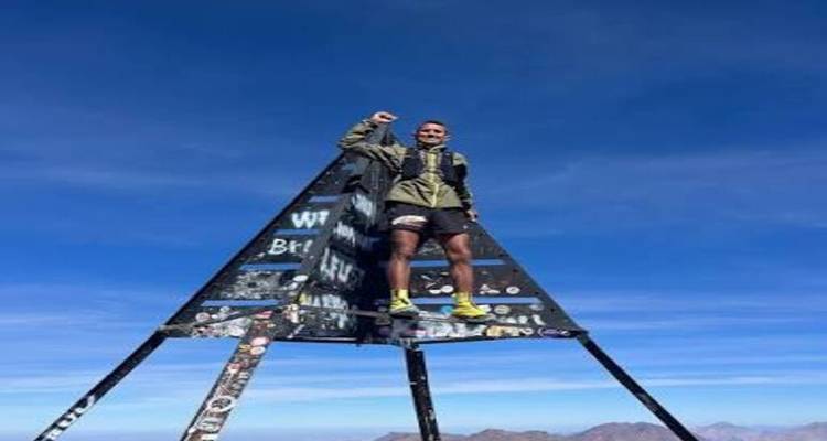 Un alpiniste célébrant au sommet de la pyramide métallique du Mont Toubkal sous un ciel bleu dégagé.