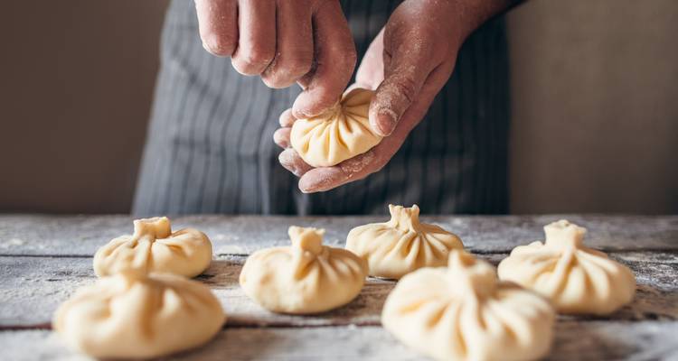 Nahaufnahme von Händen, die traditionelle georgische Khinkali-Knödel auf einem bemehlten Tisch formen.