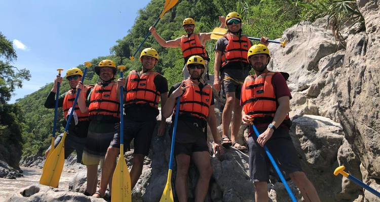 Rafting-Team, das mit Paddeln am felsigen Flussufer posiert und Sicherheitsausrüstung trägt