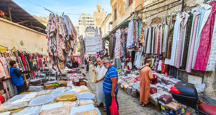 A lively market alley is lined with colorful fabrics and bustling shoppers in an Algerian city.