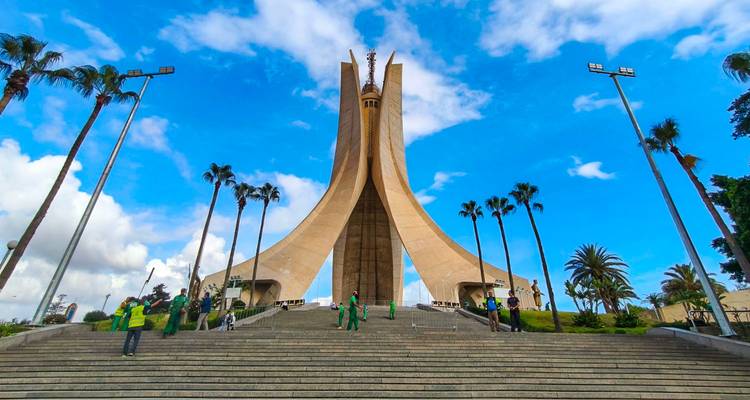 The soaring concrete wings of the Martyrs’ Memorial tower above palm-lined steps under a vivid blue sky.