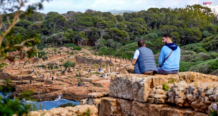 Two visitors sit on stone ruins overlooking the expansive Roman archaeological site of Tipasa beside the sea.