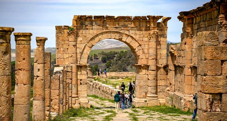 Visitors explore a grand Roman archway and columned street among ancient ruins in Algeria.