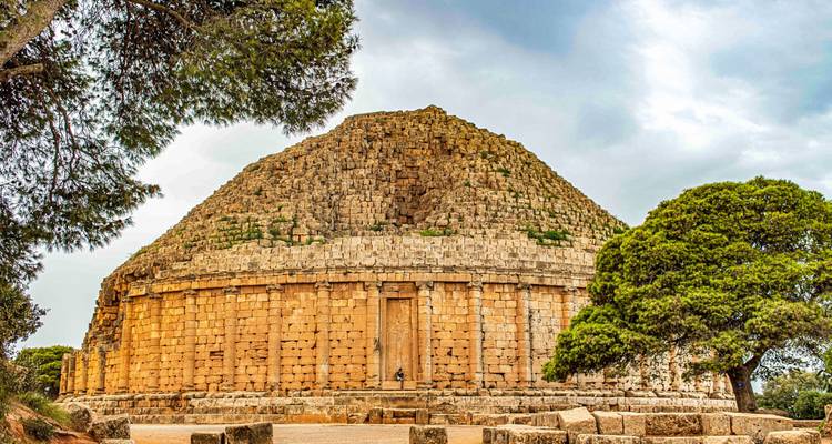 The massive stone pyramid of Medracen rises amid pines under a brooding sky.