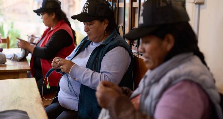 Three Andean women sit inside a workshop knitting colorful yarn with concentration.