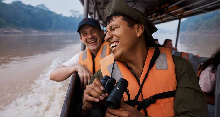 Two travellers wearing life-jackets laugh while cruising a jungle river in an open boat, one holding binoculars.